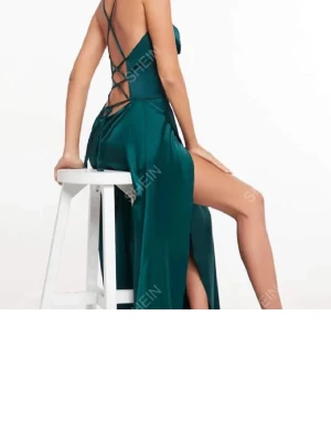 Grön långklänning med snörning  - Elegant långklänning i djupgrön färg från SHEIN. Klänningen har smala axelband med snygg snörning i ryggen och en hög slits på sidan. Tillverkad i ett glansigt material som ger en lyxig känsla. Perfekt för speciella tillfällen.