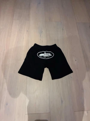 Corteiz shorts - Svarta shorts med ett stort vitt tryck framtill som föreställer ett bergsmotiv. Shortsen har en avslappnad passform och är perfekta för sommaren. De har resår i midjan och är tillverkade i ett mjukt material som känns skönt mot huden.