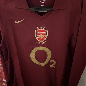 Arsenal Highbury tröja vinröd Nike - Vinröd långärmad Arsenal Highbury 1913-2006 tröja från Nike med guldfärgade detaljer och O2-logga på bröstet. Broderat Arsenal-emblem och klassisk krage. Perfekt för fotbollsfans som vill sticka ut på läktaren.