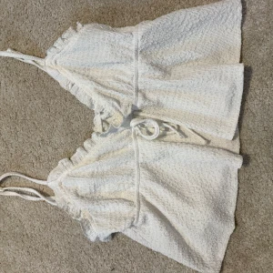 Vit omlott linnetopp från SHEIN - Säljer en söt vit omlott linnetopp från SHEIN med tunna axelband och volangdetaljer längs kanterna. Toppen har knyt i midjan och är tillverkad i ett strukturerat tyg som känns luftigt och perfekt för varma dagar.