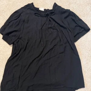 Svart blus med knytband i halsen - En svart blus med korta ärmar och knytband vid halsringningen. Blusen har en lös passform och är tillverkad i ett lätt, strukturerat tyg med diskret prickmönster. Perfekt för en avslappnad men ändå stilren look.