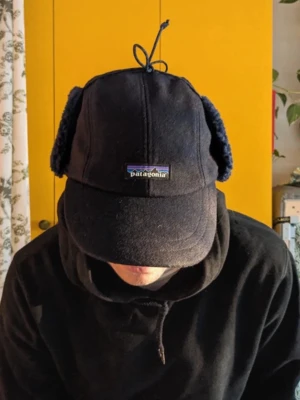 Svart fleecekeps från Patagonia - !!Bilderna är tagna av han jag köpte den av, priset kan diskuteras. Mysig svart keps från Patagonia i fleece med öronlappar och snörning upptill. Perfekt för kalla dagar när du vill hålla både huvud och öron varma. Klassisk böjd skärm och diskret logga framtill.