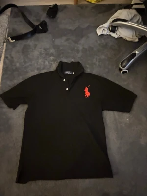 Svart pikétröja från Ralph Lauren - Klassisk svart pikétröja från Ralph Lauren med stor röd logga på bröstet och siffran 3 på ena ärmen. Tröjan har korta ärmar, krage och knappar framtill. Perfekt för en clean och sportig look.