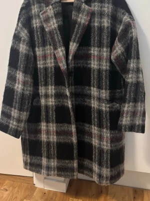 Rutig ullrock i svart och grått - Snygg rutig rock med svart, grått och diskreta röda ränder. Oversized passform med bred krage och dolda fickor på sidorna. Tillverkad i ett mjukt, ulligt material som ger en cool och avslappnad look. Perfekt för kyliga dagar.