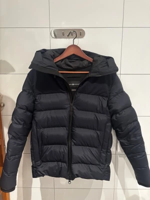 Marinblå dunjacka från Sail Racing - Säljer en Marinblå pufferjacka från Sail Racing med huva och dragkedja framtill.  Perfekt för kalla vinterdagar och även våren. Riktigt skön att bära. Jag köpte jackan på Johnells för 2-3 månader sen för 6000kr. Jag har kvittot och alla tags osv även påsen. Priset går att diskutera