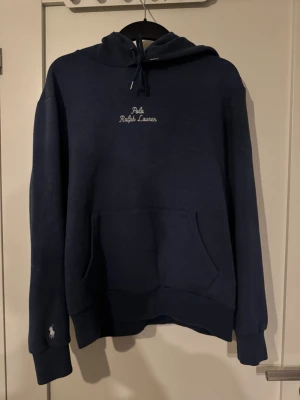 Mörkblå hoodie från Polo Ralph Lauren - Snygg mörkblå hoodie från Polo Ralph Lauren med broderad logga på bröstet och liten vit häst på ärmen. Klassisk känguruficka framtill, ribbade muddar och justerbar huva med snören. Perfekt för en chill och stilren look. Hoodien är i storlek M men passar mer någon med storlek S. Köpt ny på NK i Stockholm i somras och säljs då den inte passar mig längre.