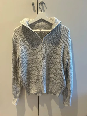 Grå stickad tröja med halvzip - Mysig gråmelerad stickad tröja med halvzip och hög krage. Tröjan har långa ärmar, ribbstickade muddar och en avslappnad passform. Perfekt för lager på lager och enkel att styla till jeans eller kjol.