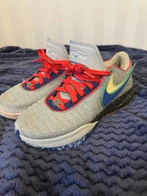 Nike LeBron 20 med färgglada detaljer - Snygga basketskor från Nike med grå mesh-överdel, röda snören och blå samt gula detaljer på sidan. Skorna har en svart sula med vita prickar och färgade inslag. Insidan är ljusgrå och kanten har röda accenter. Perfekta för dig som gillar bra och snygga basketskor.