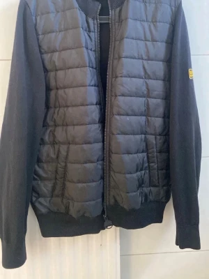 Svart pufferjacka från Gant - Snygg svart pufferjacka från Gant med quiltad front och stickade ärmar samt rygg. Jackan har ribbade muddar och dragkedja framtill. Liten Gant-logga på ärmen. Perfekt för kalla vinterdagar och riktigt stilren look.