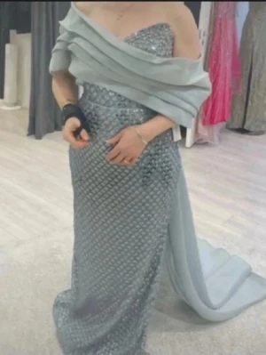 Lång glittrig blå balklänning - Elegant långklänning i glittrigt blått tyg med offshoulder-design och draperad överdel. Klänningen har en figurnära passform och ett vackert fallande släp. Perfekt för bal eller festliga tillfällen.