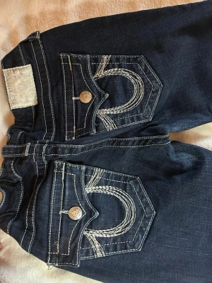 Mörkblå True Religion jeans - Säljer ett par mörkblå jeans från True Religion med  stora fickor bak med snygga detaljer. Klassisk rak passform och coola metallknappar. Det är storlek W29 men jag har sytt och fixat midjan till en w25/26. 