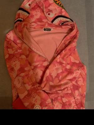 BAPE rosa camo hoodie med tigerhuva - Säljer en ikonisk rosa camo hoodie från BAPE med dragkedja och unik tigerdesign på huvan. Hoodien har ett färgstarkt mönster i olika rosa nyanser och detaljer i vitt och orange. Perfekt för dig som vill sticka ut med streetwear från ett välkänt märke.