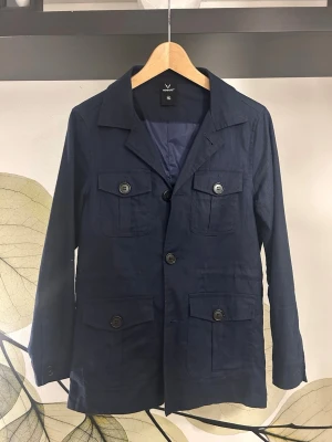 Mörkblå Kavaj från AOWOFS - Stilren mörkblå kavaj från Academy med fyra framfickor med lock och knappar. Klassisk krage och enkelknäppt design med tre knappar framtill. Tillverkad i ett lätt tyg, perfekt för en clean och modern look.