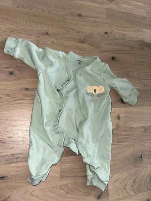 Mintgrön body med koalamotiv från Name It - Supersöt mintgrön body från Name It med tryck av en koala och texten 'lovable' på bröstet. Långärmad modell med tryckknappar framtill och i grenen för enkel påklädning. Tillverkad i mjuk bomull, perfekt för de allra minsta.