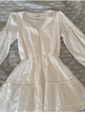Vit broderad klänning med puffärm - Söt vit klänning med broderie anglaise och v-ringning. Klänningen har långa puffärmar, markerad midja och volangkjol. Tillverkad i ett lätt bomullsliknande material med fina spetsdetaljer längs kanterna. Perfekt för sommarens alla tillfällen.