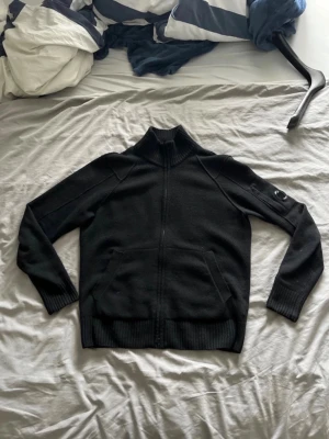 Svart ziptröja från C.P. Company - Säljer en svart ziptröja från C.P. Company med hög ribbad krage och två fickor framtill. Tröjan har en diskret logga på ärmen och är tillverkad i ett mjukt stickat material. Perfekt för dig som gillar stilrena och funktionella plagg. Plagget är ursprunget storlek S (46) men passar bättre som M