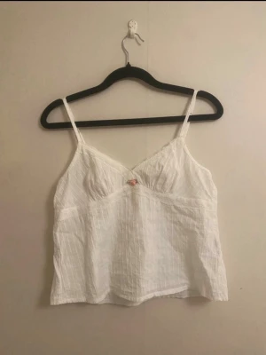 Vit linnetopp med spetsdetaljer - Söt vit linnetopp med tunna axelband och diskreta spetsdetaljer längs urringningen. Toppen har en liten rosett framtill och är tillverkad i ett lätt, luftigt tyg med subtila ränder. Perfekt för varma dagar och enkel att styla till olika looks.Vet ej vart den är ifrån köpte den på second hand och lappen är bortklippt. Skulle säga storleken är xs/s. Men vet inte då lappen är bortklippt.
