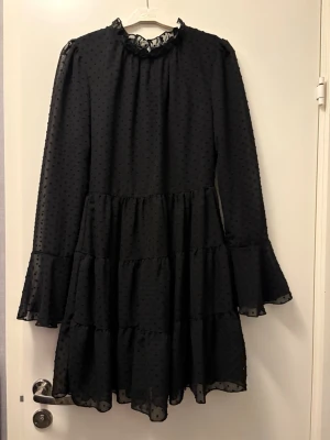 Svart volangklänning med prickar - Svart kortklänning med volanger och diskret prickigt mönster. Klänningen har långärmade ärmar med utsvängda ärmslut och hög hals med liten volangkant. Tillverkad i ett lätt, transparent material med foder under.