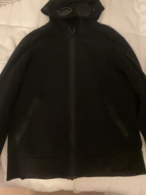 Svart cp company, vill ba få bort den - Cool svart jacka med huva och inbyggda glasögon i huvan. Jackan har dragkedja framtill och två fickor med dragkedja. Perfekt för dig som gillar streetwear och vill sticka ut lite extra.