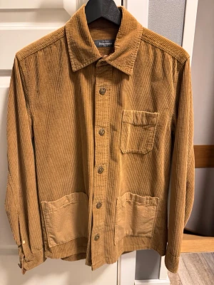 Brun manchester overshirt - Snygg brun overshirt i manchester från Knowledge Cotton Apparel. Klassisk krage, knäppning framtill och tre fickor – två stora nedtill och en bröstficka. Perfekt lager på lager-plagg med avslappnad passform och schyssta detaljer.