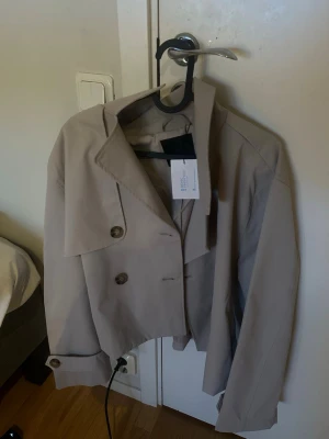 Beige trenchcoat - Stilren beige cropped trenchcoat med dubbelknäppning och bred krage. Jackan har dekorativa knappar vid ärmsluten och klassiska trenchcoat-detaljer. Perfekt för dig som vill ha en modern twist på en klassiker.