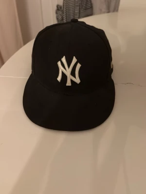 Svart New York Yankees keps - Snygg svart keps från New Era med klassiska New York Yankees-loggan broderad i vitt framtill. Kepsen har böjd skärm och MLB-logga baktill. Perfekt accessoar för en sportig och urban stil.