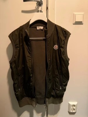 Moncler dunväst Olivgrön - Snygg olivgrön dunväst från Moncler med ribbade muddar och krage. Västen har dragkedja framtill, två fickor med dragkedja och Moncler-logga på bröstet. Perfekt för lager-på-lager och streetstyle. Storlek L men sitter mindre. Handtag är lös