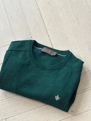 Mörkgrön Morris tröja  - Snygg mörkgrön stickad tröja från Morris med rund halsringning och diskret broderad lilja i vitt på bröstet. Tröjan har en klassisk passform och ribbade muddar vid halsen. Perfekt för dig som gillar stilrena detaljer och mjuka material.