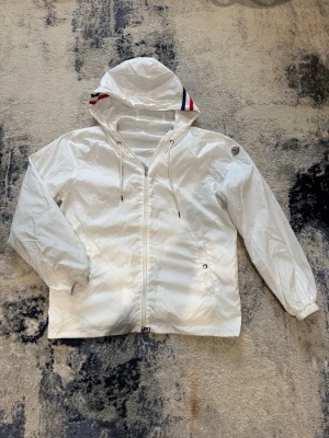 Vit vindjacka från Moncler - Snygg vit vindjacka från Moncler med huva och dragkedja framtill. Jackan har Moncler-logga på ärmen och coola rödblå ränder på huvan. Lätt material, perfekt för blåsiga dagar. Justerbar huva med snören och elastiska muddar vid ärmslut.