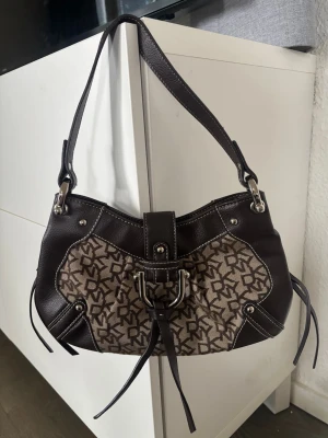 Unik Brun axelväska från DKNY - Snygg axelväska från DKNY i brunt läder och canvas med DKNY-logga över hela väskan. Väskan har silverfärgade metalldetaljer, dekorativt spänne framtill och vita kontrastsömmar. Perfekt storlek för det viktigaste och en cool detalj till din outfit. Använd gärna köp nu❤️