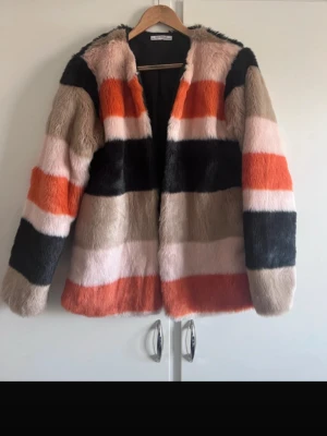 Randig fuskpälsjacka i färgblock - Säljer en fluffig fuskpälsjacka från Zara  med breda ränder i svart, beige, orange och ljusrosa. Jackan har rak passform och passar bra på både S och M, hakar för stängning och helsvart foder. Perfekt för dig som vill sticka ut med färg i vintergarderoben.