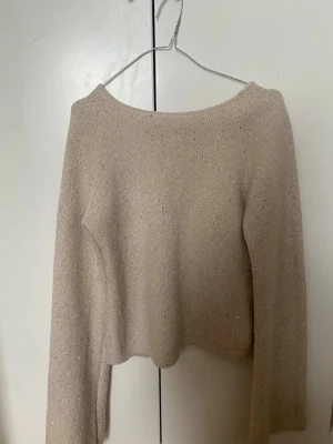 Beige stickad cardigan med knytning - Trendig beige stickad cardigan med tre knytband framtill och lätt croppad passform. Tröjan har långa ärmar och är gjord i ett mjukt, glittrigt material som ger en extra touch. Perfekt för lager på lager och snygg till både jeans och kjol.