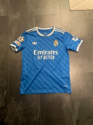 Real Madrid Bellingham matchtröja blå - Säljer en blå Real Madrid matchtröja från Adidas med vita detaljer och tryck. Tröjan har Champions League-märke på ärmen, klubbmärke och Adidas-logga på bröstet samt 'Emirates Fly Better' på framsidan. På ryggen står det Bellingham och nummer 5.