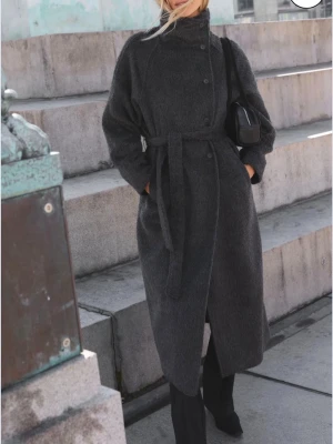 Mörkgrå ullkappa med knytband - Stilren mörkgrå kappa i ullmix med hög krage och knappar framtill. Kappan har ett matchande knytband i midjan och raka ärmar. Lång modell som passar perfekt till höst och vinter. Enkel men trendig design som funkar till många looks.Pris kan deikusteras men köpte för 600