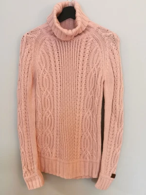 Jättefin ljusrosa polotröja med flätmönster, från Boomerang - Stickad ljusrosa polotröja med grovt flätmönster framtill och på ärmarna. Tröjan har hög polokrage, ribbstickade muddar och nederkant. Perfekt för kalla dagar och ger en mysig, trendig look. Materialet ser ut att vara mjukt och värmande. Storlek XS. Från märket Boomerang. Är i fint använt skick, lite nopprig under armarna,i övrigt fint skick. 
