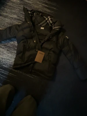 Svart Burberry pufferjscka - Snygg svart Burberry pufferjacka med huva och rutigt innerfoder. Jackan har dragkedja och knappar framtill samt två stora fickor. Perfekt för kalla dagar och har en modern, boxig passform.