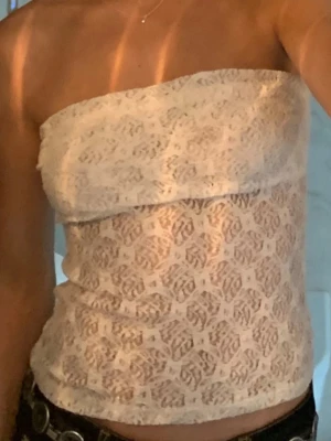Vit spets bandeau tubtopp - Säljer en snygg vit bandeau tubtopp i spets med transparent mönster. Toppen är axelbandslös och har en croppad passform som passar perfekt till höga jeans eller kjol. Perfekt för dig som vill ha en trendig och luftig topp till sommaren.