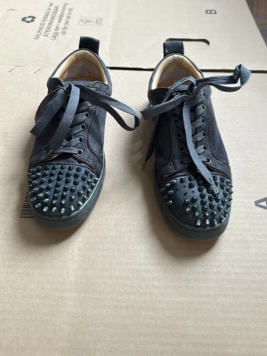 Svarta loubs - Unika svarta sneakers från Christian Louboutin med coola nitar på tån och klassisk röd sula. Skorna har svarta skosnören och är tillverkade i mocka och läder. Perfekta för dig som vill sticka ut med din stil.