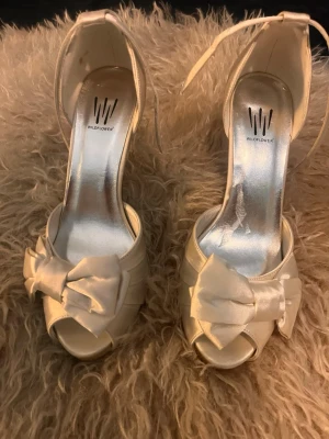 Vita pumps med rosett från Wildflower - Snygga vita pumps från Wildflower med öppen tå, smal klack och dekorativa stora rosetter framtill. Skorna har en elegant satinfinish och justerbara ankelremmar för extra komfort och stil. Perfekta för att lyfta din outfit till nästa nivå.