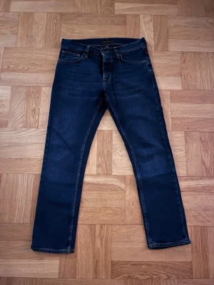 Nudie Jeans Grim Tim W30 L28– Mörkblå – Mycket fint skick - Säljer ett par Nudie Jeans modell Grim Tim i mörkblå tvätt. Klassisk slim straight-passform med normal midja och rak, smal silhuett.  Storlek: W30 L28 Färg: Mörkblå med lätt naturlig fading Skick: Mycket fint skick, inga hål eller större slitage.  Tillverkade i kvalitetsdenim med klassiska Nudie-detaljer och orange sömmar. Perfekta både till vardag och mer uppklätt med skjorta eller kavaj.  Hör av dig vid frågor eller för fler bilder 🙂