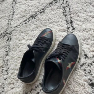 Arigato clean 90 bird - Svarta sneakers i skinn från Axel Arigato med broderade fåglar i rött, blått och beige på sidorna och tån. Klassisk vit sula och svarta skosnören. Perfekta för dig som vill ha en stilren men ändå unik look.