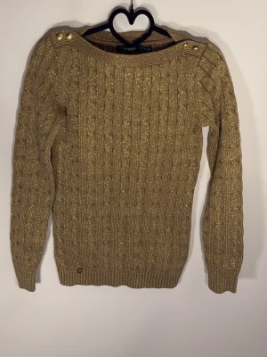 Guldig stickad tröja från Ralph Lauren - Snygg guldig stickad tröja från Lauren Ralph Lauren med kabelstickat mönster och dekorativa knappar på axlarna. Tröjan har långa ärmar och ribbade muddar. Perfekt för dig som vill sticka ut med lite extra glitter i vardagen.