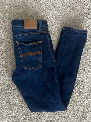 Mörkblå plate jeans från Nudie Jeans - ‼️Jeansen passar någon från 175-185 som väger 55-70kg! Säljer ett par mörkblå slim fit jeans från Nudie Jeans med klassiska orange sömmar och läderpatch bak. Jeansen har fem fickor och är tillverkade i mjuk denim med stretch. Perfekta för dig som gillar en clean och tidlös look. 