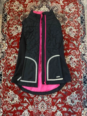 Svart dunväst med rosa detaljer  - Snygg svart dunväst från Lindex med rosa innerfoder och dragkedja. Västen har två stora fickor framtill med grå kant och reflekterande detaljer. Perfekt för lager-på-lager och sportiga outfits. Materialet är lätt och quiltat för extra värme.