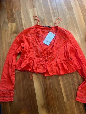 Röd croppad blus Zara - Trendig röd croppad blus från Zara med volangkant, spetsdetaljer och knyt framtill. Blusen har lång ärm och v-ringad halsringning, perfekt för dig som vill ha en färgstark och unik topp till din outfit.