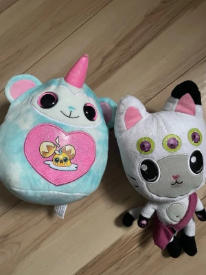 Gabbys  Dockhouse gosedjur - Gabby Dockhouse två mjuka gosedjur: en blå och rosa enhörningsbjörn med hjärtmotiv och stora ögon, samt en vit och grå katt med tre ögon, svarta öron och rosa detaljer. Perfekta som samlarobjekt för dig som gillar unika mjukdjur.