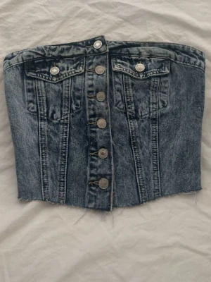 Mörkblå denim tubtopp med knappar - Snygg mörkblå tubtopp i denim med silverfärgade knappar framtill och två bröstfickor. Toppen har en bandeauform och smockad rygg för extra komfort. Rå kant nedtill ger en edgy vibe. Perfekt för dig som vill sticka ut.