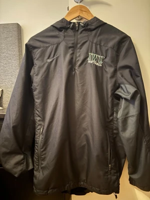 Svart vindjacka Nike IWU Softball - Svart vindjacka från Nike med IWU Softball-logga på bröstet och dragkedjor vid fickorna. Jackan har huva, halv dragkedja framtill och meshfoder för extra komfort. Perfekt för blåsiga dagar och sportiga outfits.
