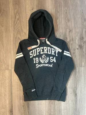 Mörkgrå Superdry hoodie med tryck - Mörkgrå hoodie från Superdry med vit text och tryck framtill, samt vita ränder på ärmarna🤩 Passar S-Xs inga stora fel förutom det som syns på sista bilden😇 hör av er vid funderingar samt snabbaffär🙌🏻