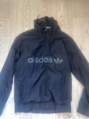 Svart vindjacka från Adidas - Svart vindjacka från Adidas med stor logga och trefoil på bröstet. Jackan har halv dragkedja, huva med dragsko och två fickor framtill. Tillverkad i lätt och slitstarkt material, perfekt för blåsiga dagar.